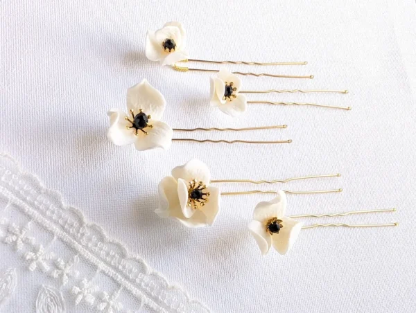 Set d'épingles à chignon pour le mariage posés à plat à côté d'un voile de mariée. Pics à chignon ornés de petites fleurs de porcelaine pésentant dans leur coeur un cristal noir et des étamines dorées