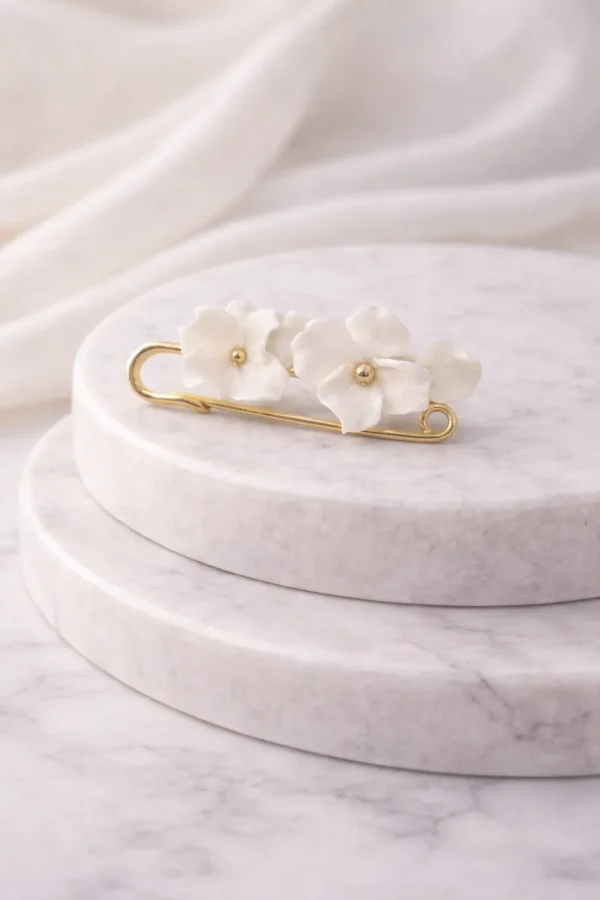 Attache-traîne DIANE 2 <p>Une épingle dorée et quelques fleurs blanches en porcelaine froide : DIANE est une attache-traîne mariée qui relève la traîne après la cérémonie, pour danser librement. Elle sert ensuite de broche ou d'attache de boléro.</p><ul><li>Fait main au Pays basque, en France</li><li>Fleurs en porcelaine froide, épingle en laiton</li><li>Longueur : 5 cm</li></ul> Attache-traîne DIANE posée à l'horizontale : fleurs en porcelaine froide sur une épingle dorée
