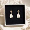 Boucles d’oreilles mariage Kate présentées dans un écrin blanc, avec goutte en porcelaine froide et puce dorée ornée de cristaux, sur fond textile délicat.