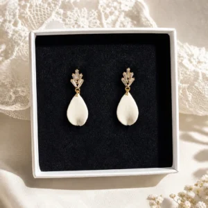 Boucles d’oreilles mariage Kate présentées dans un écrin blanc, avec goutte en porcelaine froide et puce dorée ornée de cristaux, sur fond textile délicat.