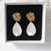 Boucles d'oreilles ZÉLIE dans leur écrin noir : puces soleil dorées et pétales blancs en porcelaine froide