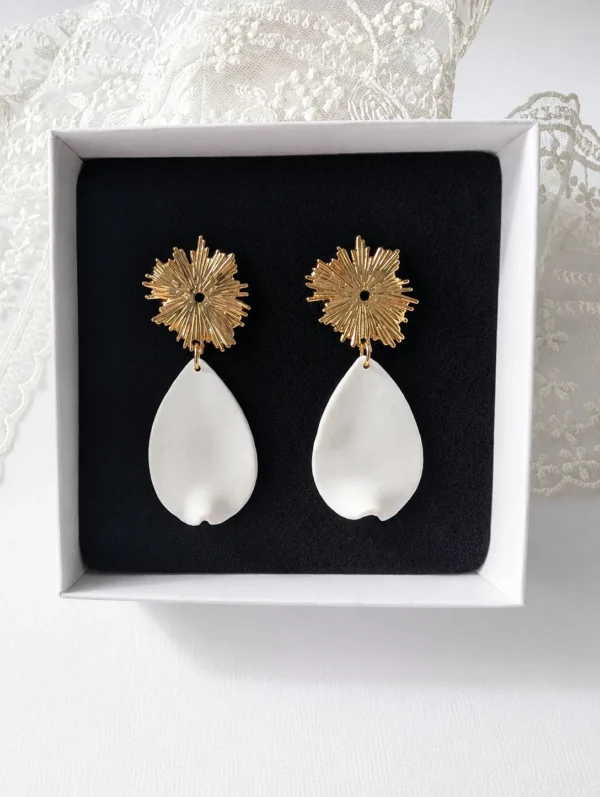 Boucles d'oreilles ZÉLIE 3 <p>Zélie associe une puce dorée en forme de soleil et un pétale blanc en porcelaine froide, à la forme libre. Des boucles d'oreilles lumineuses et légères, loin du bijou standardisé.</p><ul><li>Faites main dans mon atelier du Pays Basque</li><li>Puce soleil en laiton plaqué or 18k, pétale en porcelaine froide</li><li>Pétale de 2 × 3,3 cm · 1,8 g</li></ul> Boucles d'oreilles ZÉLIE dans leur écrin noir : puces soleil dorées et pétales blancs en porcelaine froide