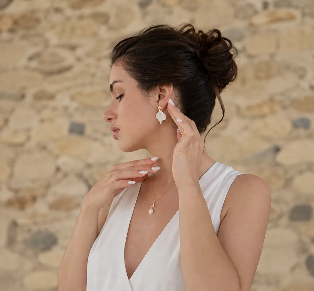 Accueil 3 Femme de profil devant un mur de pierre, chignon et haut blanc à col en V, portant les boucles d'oreilles NOÉMIE : un pétale de porcelaine froide sous une puce fleur à cristaux, by Marguerite