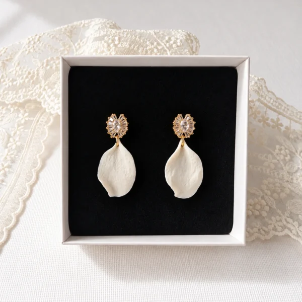 Boucles d'oreilles STELLA 11 Un pétale de porcelaine froide façonné à la main, à l'aspect de pétale de rose, sous une puce sertie de zircones cubiques : STELLA, ce sont des boucles d'oreilles de mariée à pétale de porcelaine, lumineuses et légères, en version dorée ou argentée. <ul> <li>Faites main au Pays basque, en France</li> <li>Pétale en porcelaine froide, puce plaqué or ou plaqué platine et zircones cubiques</li> <li>Pétale d'environ 3 cm · environ 1,7 g par boucle</li> </ul> Boucles d'oreilles STELLA : pétale de porcelaine froide blanc, aspect pétale de rose, sous une puce dorée sertie de zircones, dans leur écrin, by Marguerite