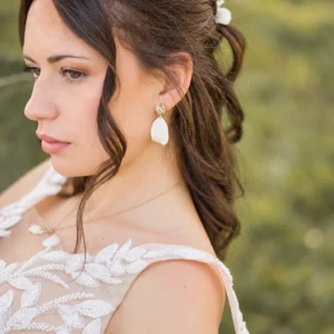 Mariée aux cheveux bruns ondulés portant les boucles d'oreilles STELLA : pétale blanc en porcelaine froide sous une puce dorée scintillante, by Marguerite