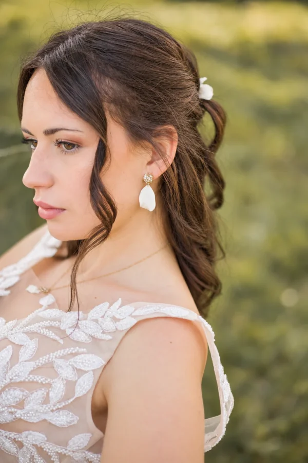 Boucles d'oreilles STELLA 12 Un pétale de porcelaine froide façonné à la main, à l'aspect de pétale de rose, sous une puce sertie de zircones cubiques : STELLA, ce sont des boucles d'oreilles de mariée à pétale de porcelaine, lumineuses et légères, en version dorée ou argentée. <ul> <li>Faites main au Pays basque, en France</li> <li>Pétale en porcelaine froide, puce plaqué or ou plaqué platine et zircones cubiques</li> <li>Pétale d'environ 3 cm · environ 1,7 g par boucle</li> </ul> Mariée aux cheveux bruns ondulés portant les boucles d'oreilles STELLA : pétale blanc en porcelaine froide sous une puce dorée scintillante, by Marguerite