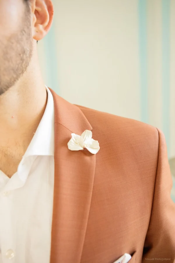 Boutonnière VIVIENNE 4 <p>Trois pétales de porcelaine froide nacrée, striés et légèrement en mouvement, avec de toutes petites perles naturelles en leur cœur : VIVIENNE est une boutonnière nacrée pour le marié. Elle répond à toute la ligne VIVIENNE de la mariée, de la couronne à l’attache-traîne.</p><ul><li>Fait main au Pays basque, en France</li><li>Pétales en porcelaine froide nacrée, petites perles naturelles</li><li>Fleur d’environ 4,8 cm de large et 4 cm de haut · épingle argentée</li></ul> Boutonnière pour témoin de mariage VIVIENNE piquée au revers d’une veste terracotta, fleur nacrée, by Marguerite