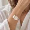 Bracelet JUSTINE porté au poignet avec une chemise blanche en gaze de coton, trois pétales blanc mat sur chaîne dorée, by Marguerite