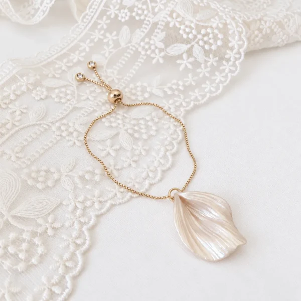 Bracelet VIVIENNE 3 Un seul pétale de porcelaine froide nacrée, façonné et texturé de fines stries, au bout d’une chaîne dorée réglable : VIVIENNE est un bracelet pétale nacré pour la mariée. Elle le garde ensuite au poignet, au quotidien. <ul> <li>Fait main au Pays basque, en France</li> <li>Pétale en porcelaine froide nacrée, environ 3 cm de long et 2,5 cm de large</li> <li>Chaîne dorée réglable, un petit zirconium à chaque extrémité</li> </ul> Bracelet VIVIENNE posé sur une dentelle : chaîne réglable dorée, curseur et petits zirconiums aux extrémités, by Marguerite