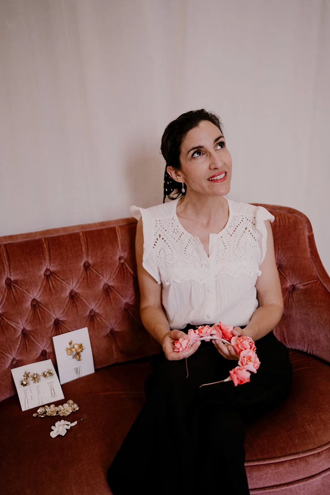 Accueil 16 Céline, créatrice de by Marguerite, assise sur un canapé de velours rose, une couronne de fleurs de porcelaine corail entre les mains, à côté de ses épingles à cheveux dorées, by Marguerite