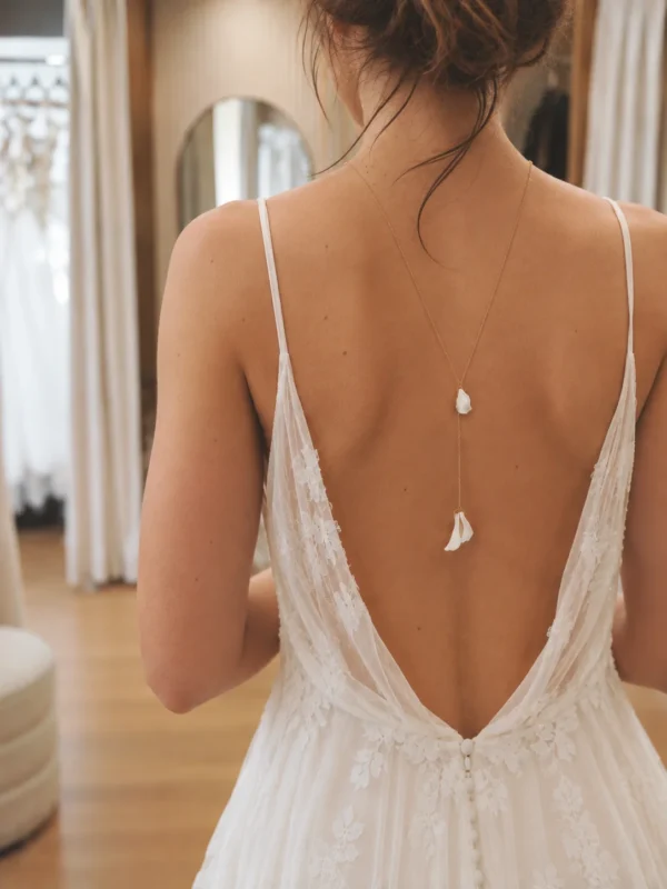 Collier de dos DIANE 8 Une perle ronde à l'avant, et dans le dos trois petits pétales de porcelaine qui bougent au moindre pas : DIANE est un collier de dos de mariage aux pétales de porcelaine, léger et délicat. Sa chaîne fine dessine une ligne de lumière le long d'une robe dos nu, du jour J aux soirées d'été. <ul> <li>Fait main au Pays basque, en France</li> <li>Chaîne gold filled 14 carats, perle de culture, pétales en porcelaine froide</li> <li>Longueur 65 ou 70 cm, pendant de 10 cm · sur mesure possible</li> </ul> Collier de dos DIANE porté avec une robe de mariée dos nu en dentelle, pétales blancs le long du dos, by Marguerite