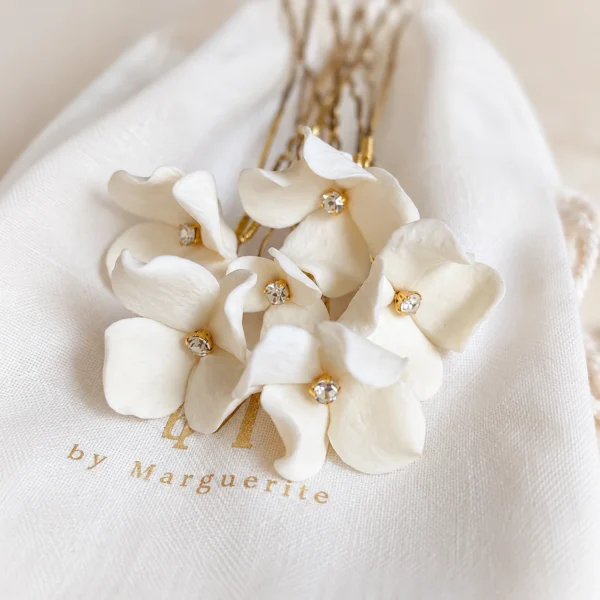 Épingles PAULINE 5 <p>Une petite fleur de porcelaine froide à peine éclose par épingle, chacune différente, de 1,5 à 3 cm, autour d’un strass : PAULINE, ce sont des épingles à chignon de mariée que vous choisissez à l’unité, de deux à sept. Je compose l’assortiment en mêlant les tailles, pour que les fleurs s’accordent comme un petit bouquet dans votre coiffure.</p><ul><li>Fait main au Pays basque, en France</li><li>Fleurs en porcelaine froide, cœur en strass, épingles en U de 7 cm</li><li>De 2 à 7 épingles · version dorée ou argentée</li></ul> Épingles PAULINE dorées posées sur un pochon en lin : six fleurs de porcelaine froide au cœur scintillant, by Marguerite