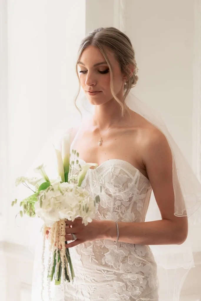 Mariée en robe de dentelle et voile, bouquet blanc à la main, dans une pièce lumineuse, by Marguerite