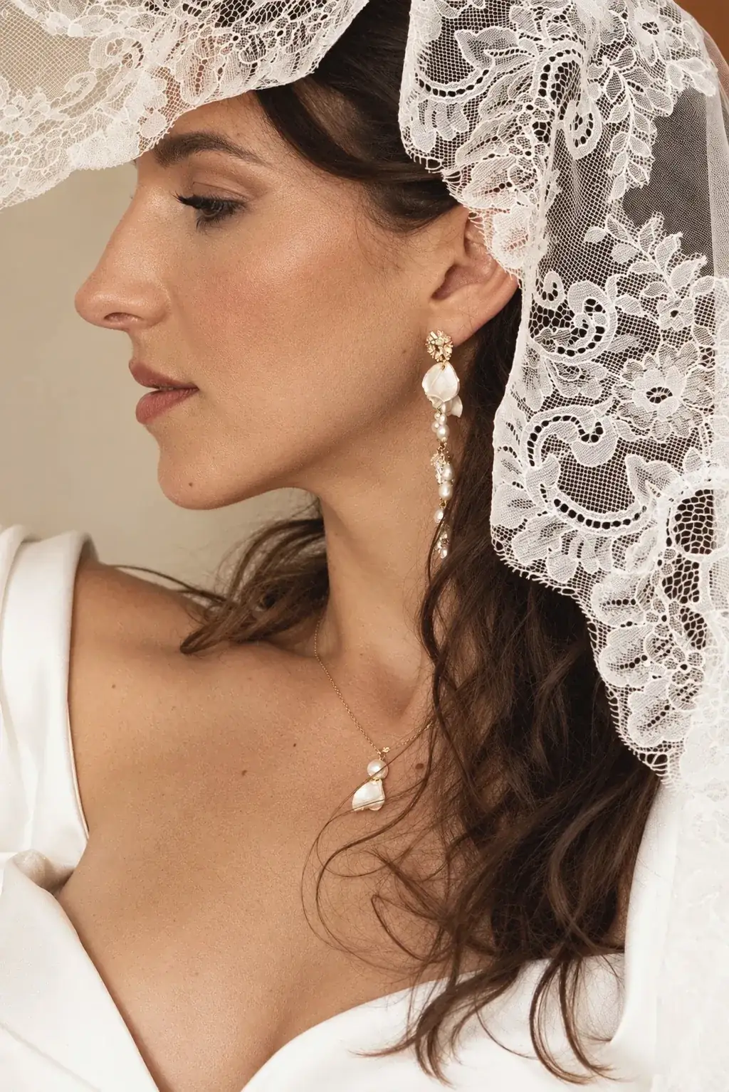 Accueil 1 Mariée de profil sous un voile de dentelle, portant les longues boucles d'oreilles ADÉLAÏDE en pétales de porcelaine froide et perles, et le collier VIVIENNE assorti, by Marguerite